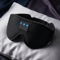 Black wireless eye mask with built-in control buttons, resting on a white pillow with a dark blanket in the background.