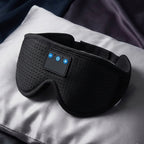 Black wireless eye mask with built-in control buttons, resting on a white pillow with a dark blanket in the background.