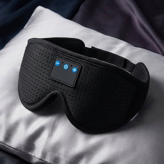 Black wireless eye mask with built-in control buttons, resting on a white pillow with a dark blanket in the background.